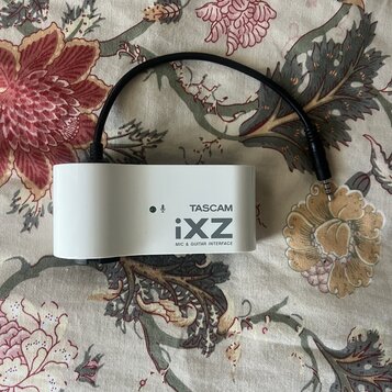 Bílé obdélníkové zařízení s připojeným černým kabelem zakončeným jack konektorem. Na boku je uveden nápis TASCAM, model iXZ a text MIC & GUITAR INTER…