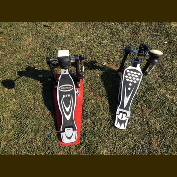 Dvě červeně a stříbrně provedená pedálová zařízení (double bass drum pedals) položená na trávě. Na červeném pedálu je viditelný nápis Millenium a pod…