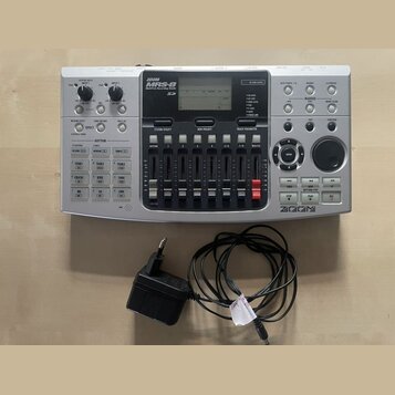 Stříbrošedý digitální nahrávací multitrack s nápisem ZOOM MRS-8 Natural Recording Studio. Zařízení obsahuje displej, ovládací knofíky, řadu posuvníků…
