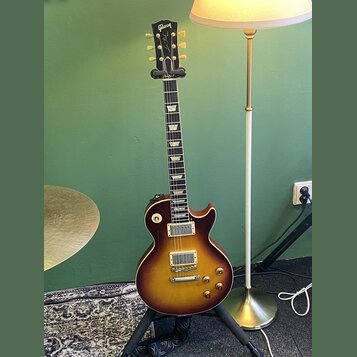 Elektrická kytara s trupem v barvě sunburst a tmavým hmatníkem s trapezoidními vložkami. Na hlavě je bílý nápis Gibson a kurzivní nápis Les Paul. Nás…