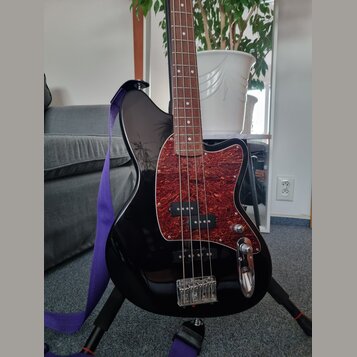 Černá elektrická basová kytara s tmavě hnědým hmatníkem. Nástroj má červený pickguard s texturou imitující dřevo a dva snímače. Na těle není vidné žá…