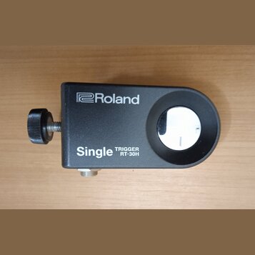 Černý trigger s nápisy Roland a Single TRIGGER RT-30H. Zařízení má obdélníkový tvar s vykrouženým otvorem a je vybaveno šroubovacím knoflíkem. Povrch…