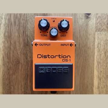 Oranžový efekt pedál s černým spínacím tlačítkem. Na zařízení jsou nápisy Distortion DS-1, BOSS, TONE, LEVEL, DIST, OUTPUT, INPUT a CHECK. Pedál má t…