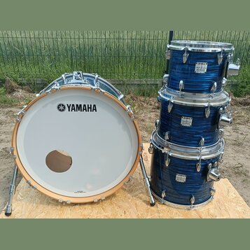 Modrá bicí souprava s kovovým hardwarem. Na bílém bendášu basového bubnu je černé logo a nápis YAMAHA. Na třech tom-tamech jsou viditelné bílé štítky…