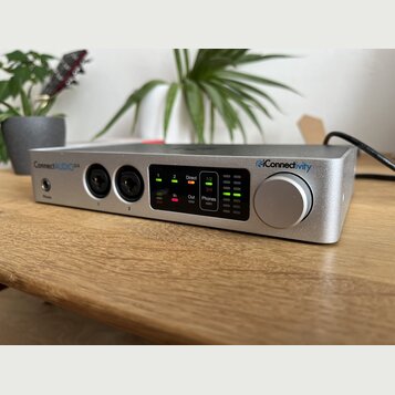 Stříbrné audio rozhraní s kovovým povrchem a velkým ovládacím knoflíkem. Na zařízení jsou nápisy ConnectAUDIO 2/4 a iConnectivity. Přední panel obsah…