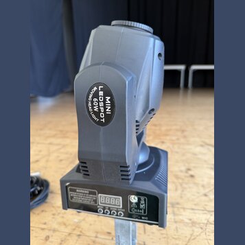 Šedý LED reflektor s pohyblivou hlavou. Na boku je černý oválný štítek s bílým nápisem MINI LEDSPOT 60W MOVING HEAD LIGHT. Spodní část obsahuje ovlád…