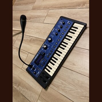 Modrý klávesový syntetizátor s bílými a černými klávesami. Na horním panelu je nápis MININOVA a logo Novation. Přístroj obsahuje několik otočných kno…