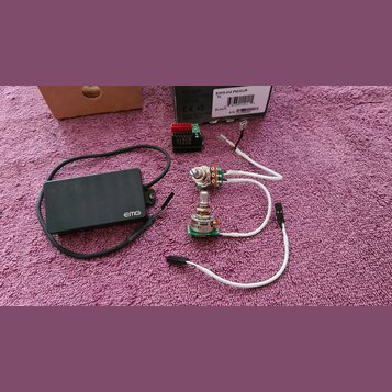 Fotografie zobrazuje sadu elektronických komponentů pro hudební nástroj. Soubor obsahuje černý obdélníkový modul s bílým nápisem EMG, dva potenciomet…