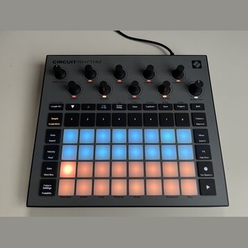 Šedý sampler s nápisem CIRCUIT RHYTHM. Zařízení obsahuje několik černých otočných ovladačů a mřížku podsvícených padů v modré a oranžové barvě. Na po…