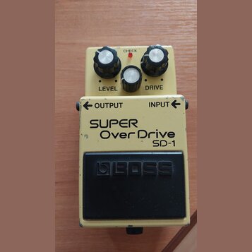 Kytarový efekt pedál v béžovém provedení s černým spínacím pedalem. Na těle jsou nápisy SUPER OverDrive SD-1, OUTPUT, INPUT a na pedálu logo BOSS. Ho…