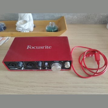 Červené audio rozhraní s nápisem Focusrite na horní straně. Na předním panelu jsou dva vstupy, ovladače zisku a knoflík s nápisy MONITOR a DIRECT MON…