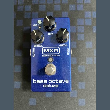 Modrý efekt pedál s nápisy MXR a BASSINNOVATIONS. Na horní části jsou tři černé ovládací knoflíky s popisky MID+, DRY, GROWL a GIRTH. Ve spodní části…