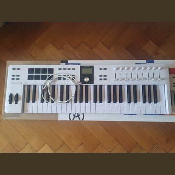 Bílá klaviatura s logem ARTURIA a nápisem KEYLAB ESSENTIAL 61. Nástroj obsahuje sadu padů, ovládací knoflíky a posuvníky. Na klaviatura leží bílý USB…