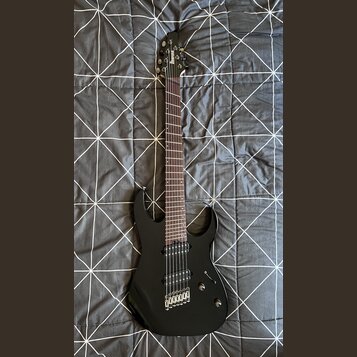 Černá elektrická kytara s tmavým hmatníkem a dvěma humbuckery. Na hlavě je bílý nápis Ibanez. Nástroj vykazuje známky běžného používání, jsou vidět d…