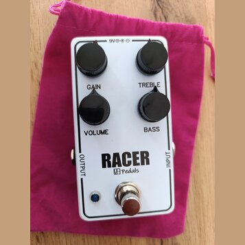 Bílý efekt pedál s nápisem RACER a logem M pedals. Na horní straně jsou čtyři černé oválné ovládací knoflíky s popisky GAIN, TREBLE, VOLUME a BASS. V…