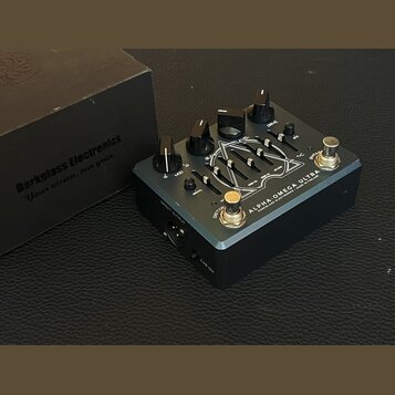Černý efekt pedál s kovovým šasi a stříbrným nápisem ALPHA·OMEGA ULTRA DARKGLASS ELECTRONICS MADE IN FINLAND. Na horní straně jsou čtyři černé otočné…