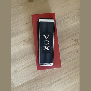 Černý kytarový wah-pedál s chromovanými okraji, umístěný na červené krabici. Uprostřed je nápis VOX a po stranách jsou texty „2 X OHMS 10W“ a „R.S.A.…