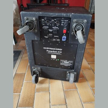 Černý aktivní subwoofer v texturovaném krytu s dvěma madly a dvěma kolečky. Na zadním panelu je ovládací sekce s konektory, potenciometry a červeným…