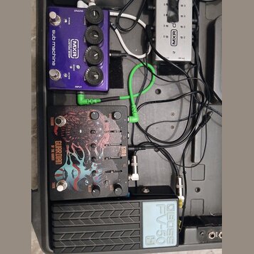 Sestava efektů v černém kufru. Obsahuje fialový pedál s nápisy MXR CUSTOM SHOP sub machine, stříbrný napájecí adaptér s logem MXR a černý pedál s náp…
