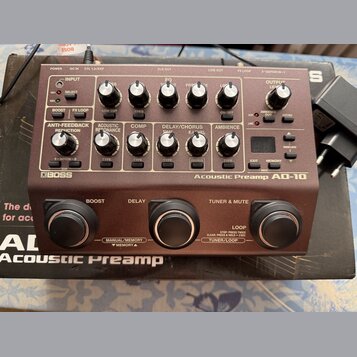 Hnědý kytarový předzesilovač s nápisy BOSS a Acoustic Preamp AD-10. Zařízení má v horní části řadu ovládacích knoflíků, přepínačů a malý digitální di…
