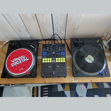Sada DJ vybavení složená ze dvou černých gramofonů a jednoho mixážního pultu na dřevěném stole. Levý gramofon má červený slipmat s nápisy VINYL DIGIT…