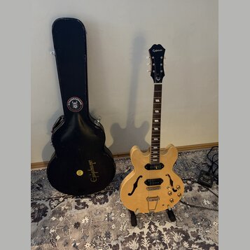 Světle hnědá polodubová elektrická kytara s dvěma humbuckery a f-otvory, umístěná na stojanu. Na hlavě hmatníku je bílý nápis Epiphone a logo v podob…