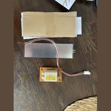 Fotografie zobrazuje elektronickou součástku s hliníkovým chladičem a přiloženou hnědou papírovou obálkou. Na oranžovém plošném spoji je bílý čtverco…