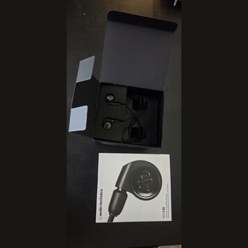 Černé špunty do uší v otevřené černé krabici. Vedle krabice je položen bílý manuál s fotografií sluchátka a nápisy audio-technica a ATH-E40. Povrchy…