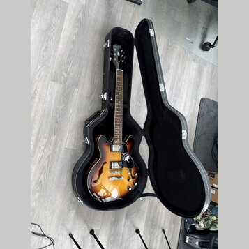 Polodupatá elektrická kytara s barevným přechodem od žluté po tmavě hnědou v černém tvrdém pouzdře. Na hlavě nástroje je bílý nápis Epiphone. Kytara…