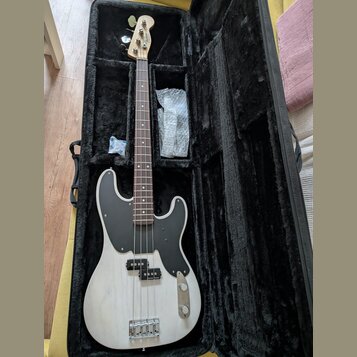 Elektrická basová kytara s bílým trupem a černým pickguardem v černém měkkém pouzdře. Na hlavě hmatníku je viditelné logo Fender. Nástroj má hnědý hm…