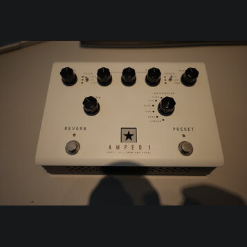 Bílý efekt pedál s nápisem AMPED 1 a podtitulem DEPT. 10 | 100W AMP PEDAL. Uprostřed je logo ve tvaru černé hvězdy v bílém čtverci. Zařízení má šest…