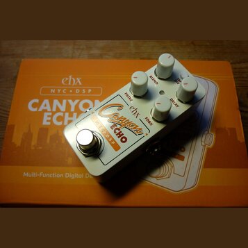 Bílý efekt pedál s nápisy ehx, Canyon ECHO a NYC DSP. Na horní straně se nachází pět bílých oválných knoflíků s popisky FILTER, BLEND, TAPE DIV, FDBK…