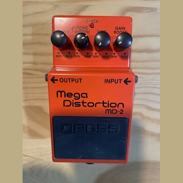 Kytarový efekt pedál oranžové barvy s modrým spínacím tlačítkem. Na přední straně je nápis Mega Distortion MD-2 a na spínači vyražené logo BOSS. V ho…