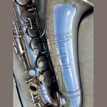 Detail stříbrného saxofonu s viditelným opotřebením povrchu a drobnými škrábanci. Na trubici jsou vyryty nápisy „H. Selmer“, „4. PLACE DANCOURT PARIS…