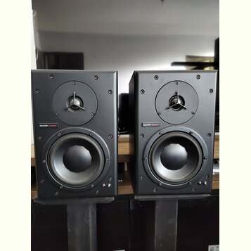Dva černé regálové reproduktory umístěné na černých stojanech. Na přední straně každého reproduktoru je nápis „dynaudio acoustics“ na červeném pozadí…