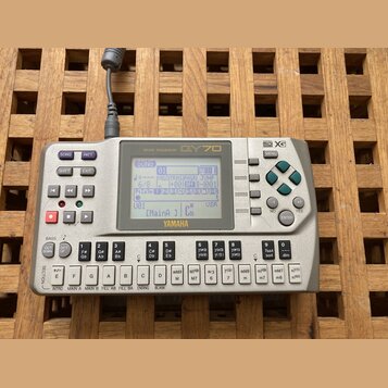 Stříbrný sekvencer Yamaha QY70 s LCD displejem. Na horní části je nápis MUSIC SEQUENCER a logo QY70, v pravém horním rohu jsou značky general MIDI a…