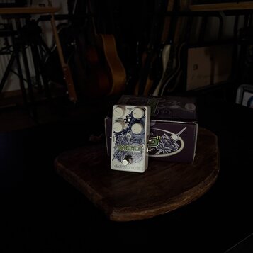 Bílý efekt pedál s nápisy MOD a electro-harmonix. Pedál má čtyři bílé ovládací knoflíky s popisky DEPTH, RATE, ATTACK a SUSTAIN. Je umístěn na dřevěn…