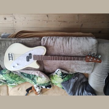 Elektrická basová kytara v krémové barvě s bílým perleťovým pickguardem. Na hlavě hnedbového krku je viditelné logo "Fender". Nástroj má čtyři struny…