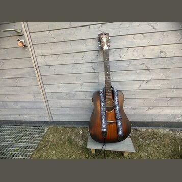 Akustická kytara s tmavě hnědým, přechodovým povrchem (sunburst) a modrým vzorovaným popruhem. Na hlavě nástroje je bílé logo Epiphone. Kytara stojí…