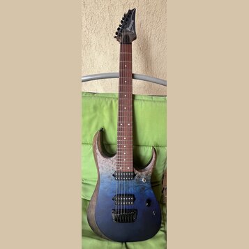Elektrická kytara s modrým průsvitným povrchem těla a viditnou strukturou dřeva. Na hlavě hmatníku je bílý nápis Ibanez. Nástroj má dva humbuckery, h…