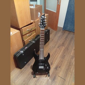 Černá elektrická kytara s špičatou hlavou a dvěma humbuckery, umístěná na stojanu. Na hlavě je bílé logo Ltd. Kytara má tmavý hmatník s bílými poloho…