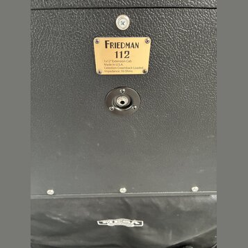 Detail zadní strany černého kytarového boxu s texturovaným povrchem. Na zlatém štítku je nápis FRIEDMAN 112, pod ním text 1x12" Extension Cab, Made i…