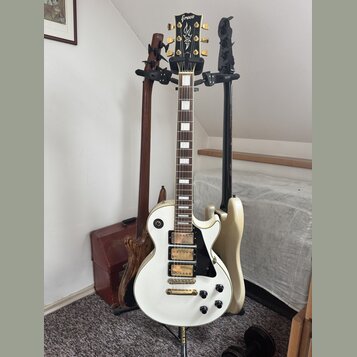 Na fotografii jsou tři elektrické kytary v držáku. V popředí je bílá kytara typu Les Paul s černým pickguardem a dvěma humbuckery, na hlavě má nápis…