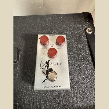 Kovový efekt pedál stříbrné barvy s třemi červenými oválnými knoflíky. Na horní části jsou nápisy MADE IN USA, 9VDC, GAIN, OUTPUT a TREBLE. Uprostřed…