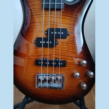 Detail těla elektrické basové kytary s lakovaným povrchem z dřeva s výraznou kresbou zrn. Nástroj má dva snímače typu humbucker v černé barvě a tři c…