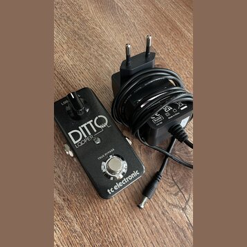 Černý kytarový pedál s nápisy DITTO LOOPER, TRUE BYPASS a tc electronic. Vedle pedálu je černý síťový adaptér s kabelem a evropskou vidlicí, na které…
