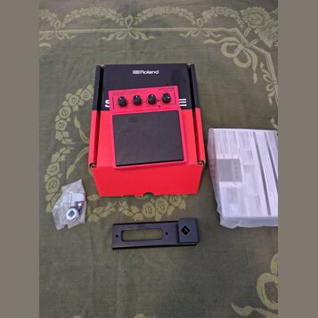 Červený elektronický sampling pad s černou gumovou plochou a nápisem Roland. Na horním ovládacím panelu je uveden model SPD-ONE WAVE PAD a několik ov…