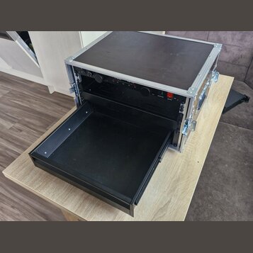 Černý transportní flightcase s hliníkovými hranami a vrškem. V horní části je osazen přístrojem s nápisy „L-Acoustics“ a „LA1I“. V dolní části je vys…