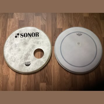 Dvě bílé bubnové kožehy položené na dřevěné podlaze. Levý kožeh s otvorem má nápis SONOR a menší štítek s textem SONOR, Natural Power. Pravý kožeh je…