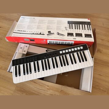 Bílá klávesová klaviatura s černým ovládacím panelem, na kterém je nápis iRig Keys I/O. Nástroj leží v otevřené krabici s polystyrenovými vložkami. V…